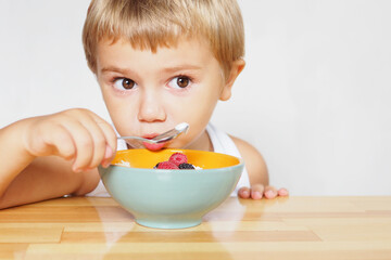 a blond boy with brown eyes in a white T-shirt eats cottage cheese with berries at a wooden table..