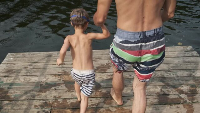 Happy family on summer vacation. Father and son having fun running on wooden pier and jumping into lake. Summertime, childhood or fatherhood concept.