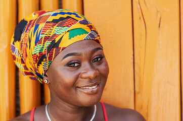African woman with a hopeful happy smile and traditional headdress standing against a painted wooden wall in the tropical village of Keta Ghana, West Africa