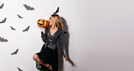 Attractive blonde girl eating halloween pumpkin. Studio portrait of inspired young woman posing in witch attire.