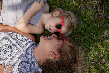 Happy little girl in red glasses is playing with her grandmother lying on the grass in the garden
