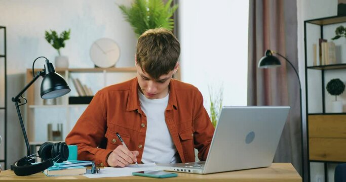 Handsome positive confident male student sitting in front of computer at home and listening online discussion from university teacher and making notes into papers