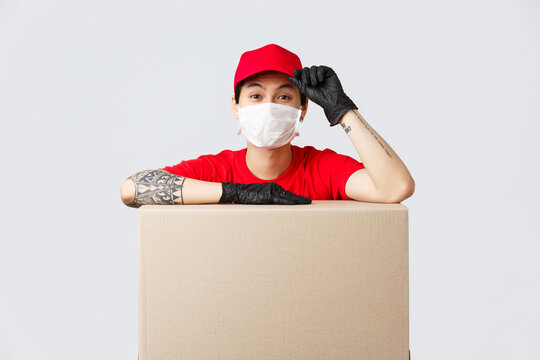 Friendly-looking asian delivery guy in red cap and t-shirt, courier service lean on cardboard box or client package, saluring as making order, transfer goods to people staying safe self-quarantine