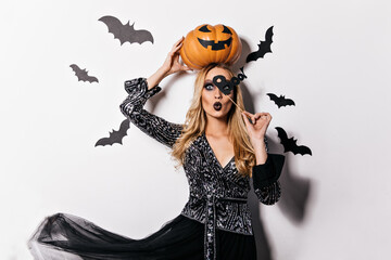 Interested long-haired girl holding orange pumpkin at halloween photoshoot. Indoor photo of appealing blonde lady in witch costume.