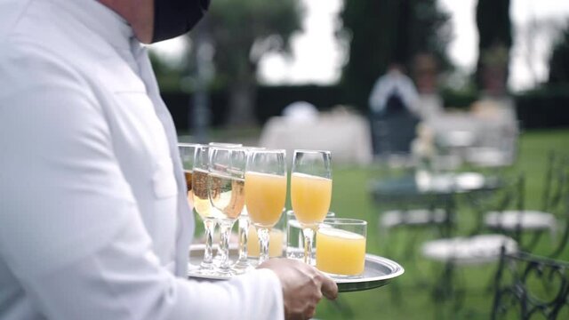 Waiter in uniform walking along the decorated tables and holding silver tray with glasses of champagne, fizzing white wine and fresh juice prepared for guests. Luxury beverages at celebration party