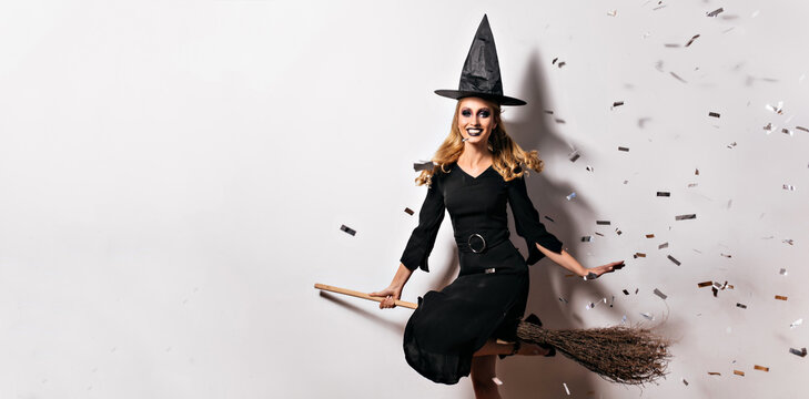 Laughing refined witch sitting on broom. Cute young wizard chilling at halloween party.