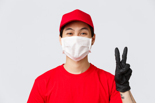 Covid-19, Self-quarantine Online Shopping Concept. Asian Courier Staying Positive, Friendly Showing Peace Sign In Medical Mask And Gloves, Measures For Safe Order Deliver On Self-quarantine