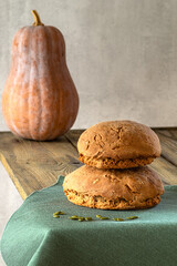 Two rolls of pumpkin bread with seeds are stacked on top of each other on a green textured napkin, there is a large, fresh pumpkin on a wooden background behind. Healthy food, trend, copy space..