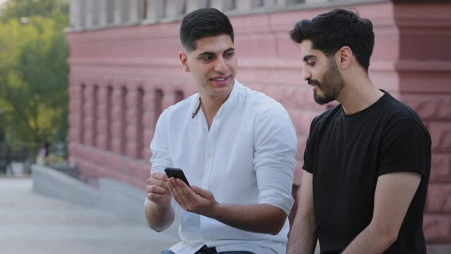 Couple of young attractive Indian Arab guys sitting outdoors, holding mobile phone, look at smartphone screen, share their impressions of vacation, show photos, watch funny video enjoying weekend