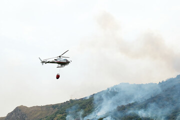 Fototapeta na wymiar Fire helicopter dropping water from a bucket on a forest fire in the mountains. Forest fire is an environmental disaster. Deforestation.