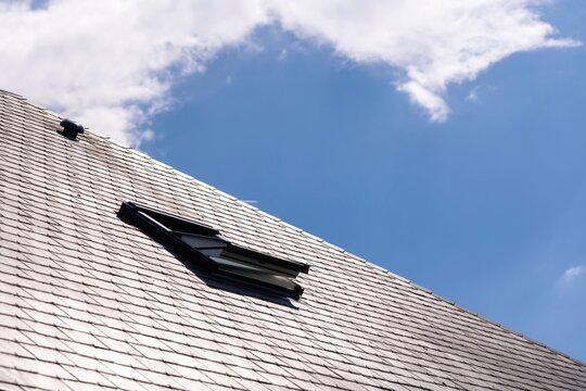 A portrait of an opened skylight window for ventilation in a slate roof on a sunny day with a blue sky with white clouds. you can also see a ventilation vent on the rooftop.