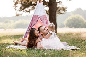 Mom, dad and little daughter are sitting next to wigwam decoration in the park. Family spending time outdoor in summer, having fun together. father's, mother's and baby's day. © Andriy Medvediuk