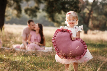 Girl are dressed in pink dress and holding big pink heart in hands. Mom, dad are sitting next to wigwam decoration in the park. Family spending time outdoor in summer, having fun together © Andriy Medvediuk