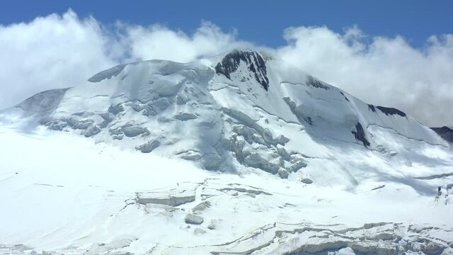 Aerial view, drone flying straight ahead. The top of the mountain is completely covered with snow against the background of blue skies and glaciers. The impregnable rock is all covered with snow