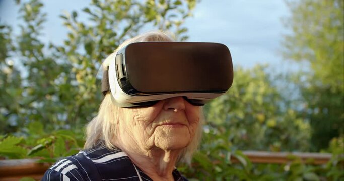 open-minded grandma turning her head in virtual reality glasses and looking at the camera smiling. pensioner raises her head to the sky in a viar helmet. positive attitude of the older generation to