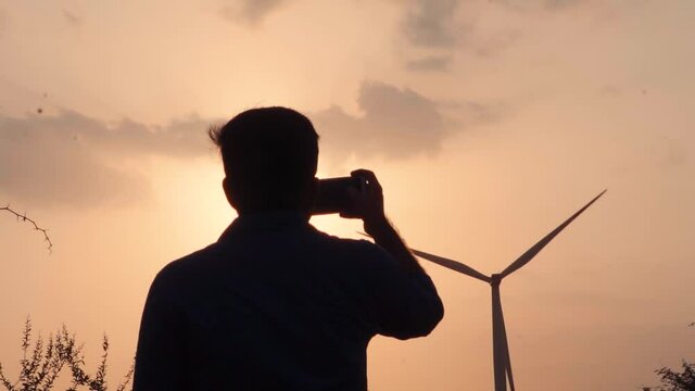 Silhouette Shot Of An Indian Tourist Taking Video Of The Sunset Besides Windmill At Wankaner In Gujarat, India. Tourist Captures Video For His Social Media. Social Media Addiction Concept. 