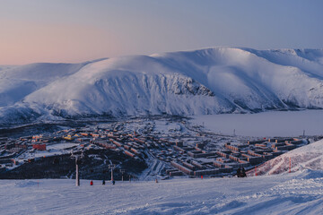 Polar city on the shore of a frozen lake surrounded by snow-capped mountains at dusk in Russia, Murmansk region, Kirovsk. Winter holiday in Arctic, Khibiny Mountains. 