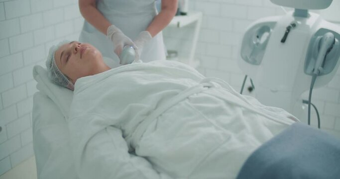 A female client is laying in the cosmetology bed in a bathrobe while a cosmetologist is doing a lifting procedure of the neck by using the Ultraformer apparatus.
