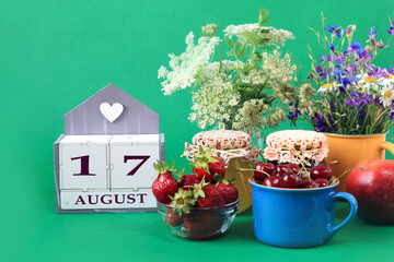 Fototapeta na wymiar Calendar for August 17 : the name of the month of August in English, cubes with the number 17, bouquets of wild flowers, jars of jam, strawberries and cherries in cups, green background
