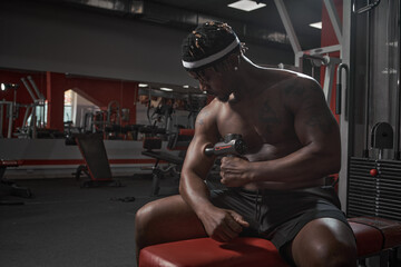 Athletic african american man massaging hand by handheld massage gun, post-workout recovery...