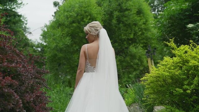 A young blonde in an elegant long white wedding dress with a long train stands with her back to the camera in a green garden, passing by delicate yellow flowers. The bride in a beautiful white dress