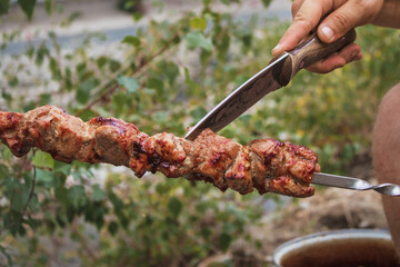 The hand of a man cutting meat fried on metal skewers with a knife. Pieces of meat are fried on a brazier. Cooking a delicious shish kebab from raw meat on coals by the fire, in the open air.