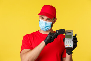 Fototapeta na wymiar Covid-19, delivery orders, shopping, contactless payment and social distancing concept. Close-up of handsome courier in red uniform and medical mask, press credit card to POS terminal
