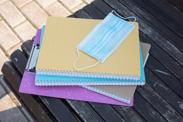 Colorful school notebooks and a protective mask lie on a bench on the street. Returning to school after the pandemic. Concept of education, health protection, medicine