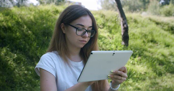 Cute teen girl in glasses working with tablet in beauty summer, spring green park. Smart, creative teenage girl writing, drawing with a stylus in a tablet. Education online, student study online