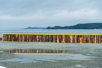 A breakwater made of wood logs and painted bright yellow on the top. There's an ocean and mountains over the wall. Before the seawater, there's a water puddle with the fence reflecting in the water.
