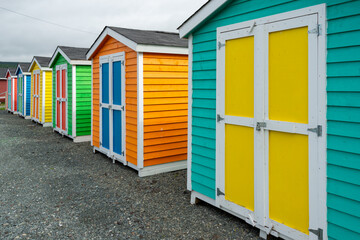 A row of small colorful painted huts or sheds made of wood. The exterior walls are colorful with double wooden doors. The sky is blue in the background and the storage units are sitting on gravel.
