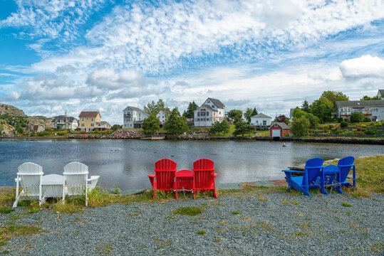 Multiple wooden Adirondack summer chairs, red, blue, and white in color along the edge of a smooth pond. The blue sky is very dramatic with colorful buildings along the edge of the water. 