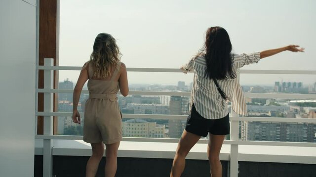 Back view of cheerful girls dancing at rooftop party doing high-five laughing enjoying view of metropolis on summer day. People and entertainment concept.