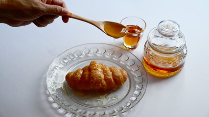 Closeup of Male hand pouring honey from a wooden spoon onto the croissant in a glass dish with honey in a glass jar and a small glass on white background. Healthy food concept, selective soft focus.