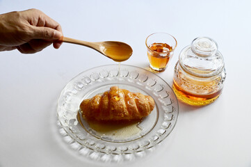 Closeup of Male hand pouring honey from a wooden spoon onto the croissant in a glass dish with honey in a glass jar and a small glass on white background. Healthy food concept, selective soft focus.