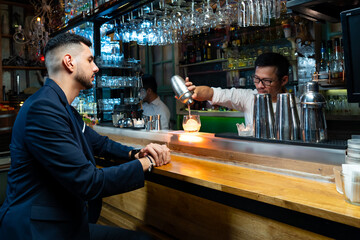 Handsome Caucasian man sitting at bar counter holding whiskey glass with ice cube and drinking alcoholic drink from barman in nightclub. Nightlife holiday party celebration and small business concept