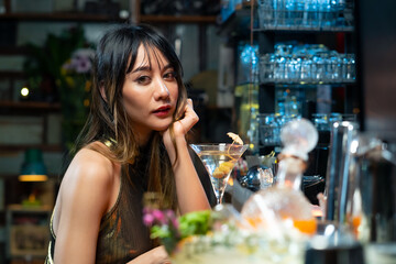 Asian woman sitting at bar counter drinking cocktail and talking to barman in nightclub. Male mixologist bartender preparing alcoholic drink to customer. Small business bar and city nightlife concept