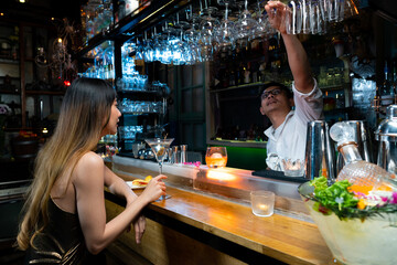 Asian woman sitting at bar counter drinking cocktail and talking to barman in nightclub. Male mixologist bartender preparing alcoholic drink to customer. Small business bar and city nightlife concept