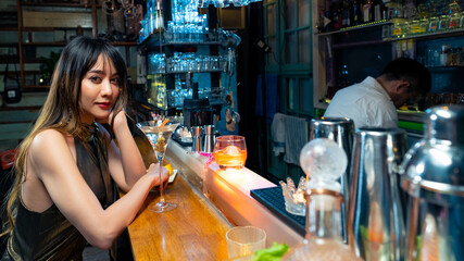 Asian woman sitting at bar counter drinking cocktail and talking to barman in nightclub. Male mixologist bartender preparing alcoholic drink to customer. Small business bar and city nightlife concept