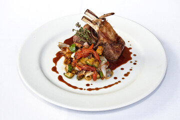 Roasted rack of lamb or rack of veal accompanied with vegetables and sauce, served on white plate over white background