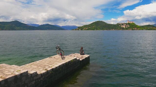 Unrecognizable solitary worried woman with bicycle behind sitting on jetty edge of Maggiore lake, Italy
