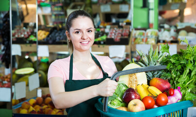 Friendly young saleswoman with shopping basket with greengrocery in grocery