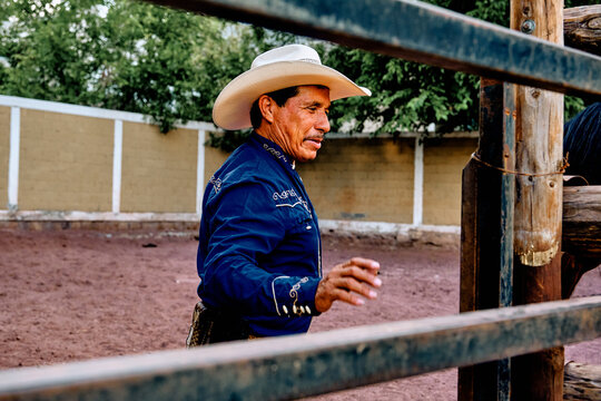 Mexican, hispanic, latino middle aged rancher in a blue shirt wearing a cowboy hat in a riding hall opening the fence for the horses