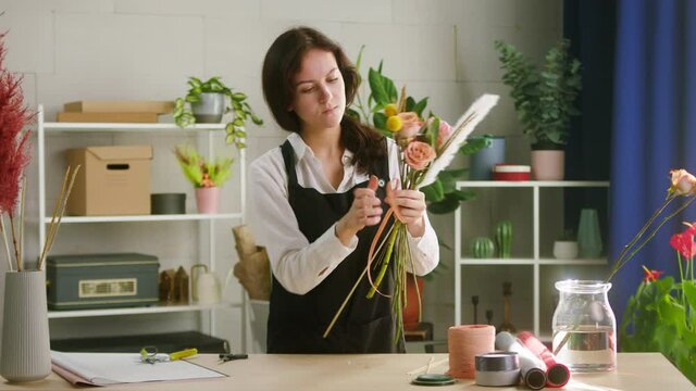 Young smiling florist making bouquet of roses and dried flowers at workplace. Floral artist working in flower shop studio. Floristry showing masterclass on creating flower arrangement. Small business.