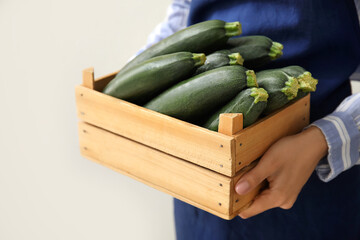 Woman holding wooden box with fresh zucchini squashes on light background, closeup