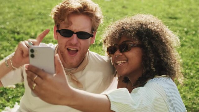 Smiling couple man and woman wearing sunglasses are taking selfie photo sitting on the grass outside
