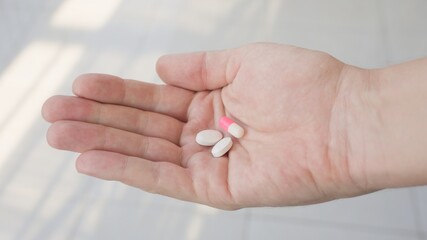 A sick person shows on the palm of his hand two white tablets and a pink capsule that the doctor prescribed for him. Against the background of a hospital white floor on a sunny day. 