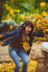 Beautiful young mother and her happy daughter are having fun in the forest at autumn. Walks in an autumn park with the kids. Family spending great time playing together with fallen leaves.