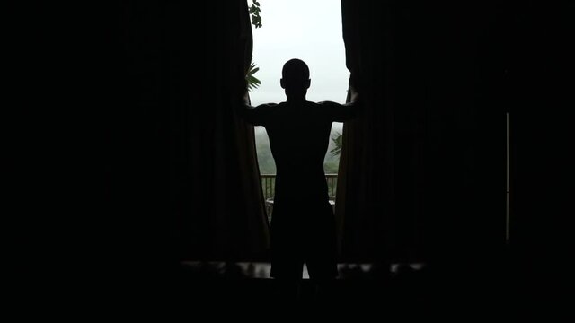Silhouetted mann opens blinds to reveal balcony in foggy rain forest, slow motion