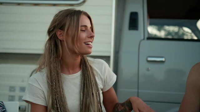 A beautiful woman is sitting and talking  in a company of friends during picnic near the caravan trailer outside